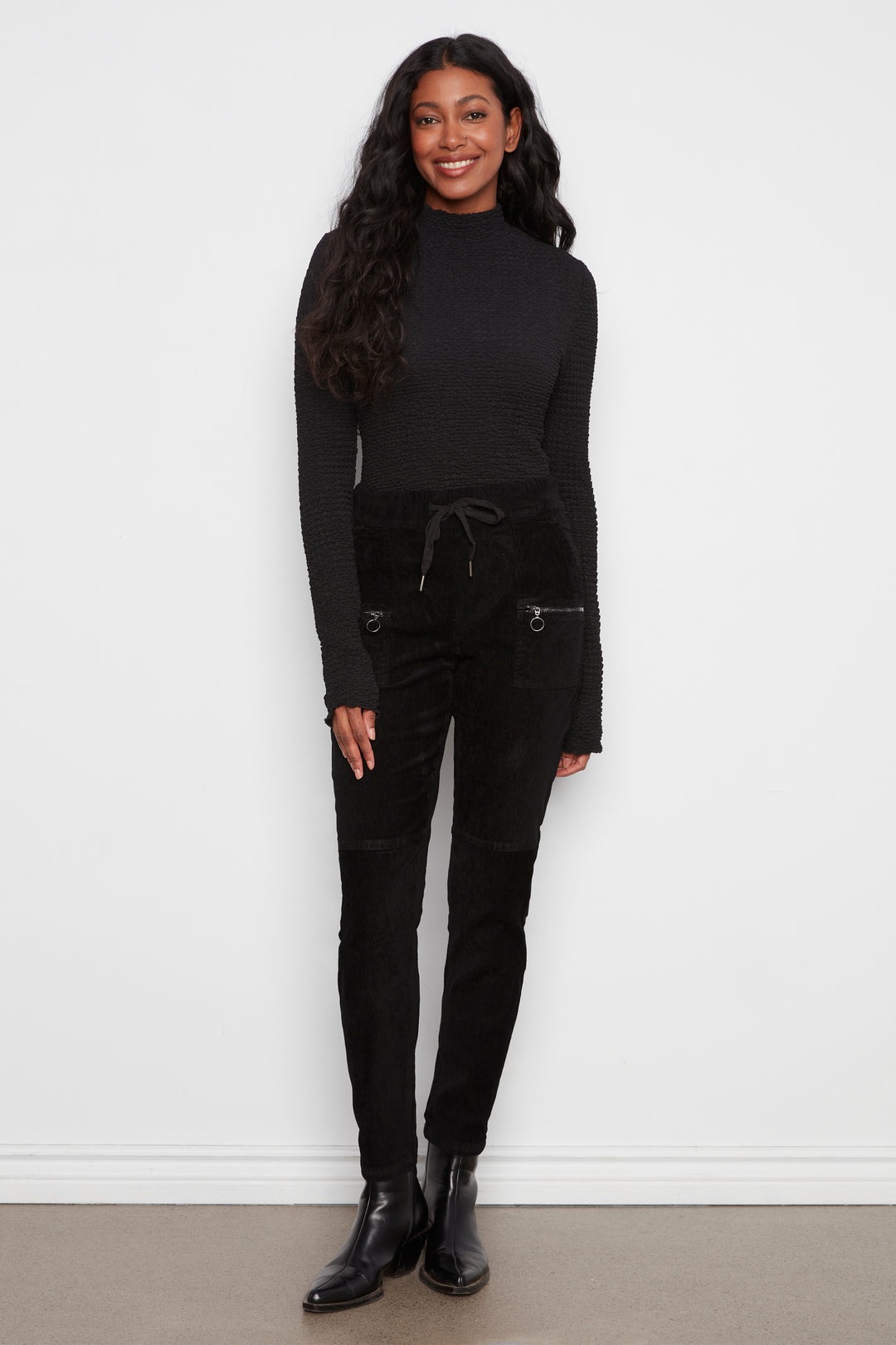 A woman modeling a black ribbed long-sleeve mock neck top and black corduroy pants with zippered pockets, standing against a plain white wall.