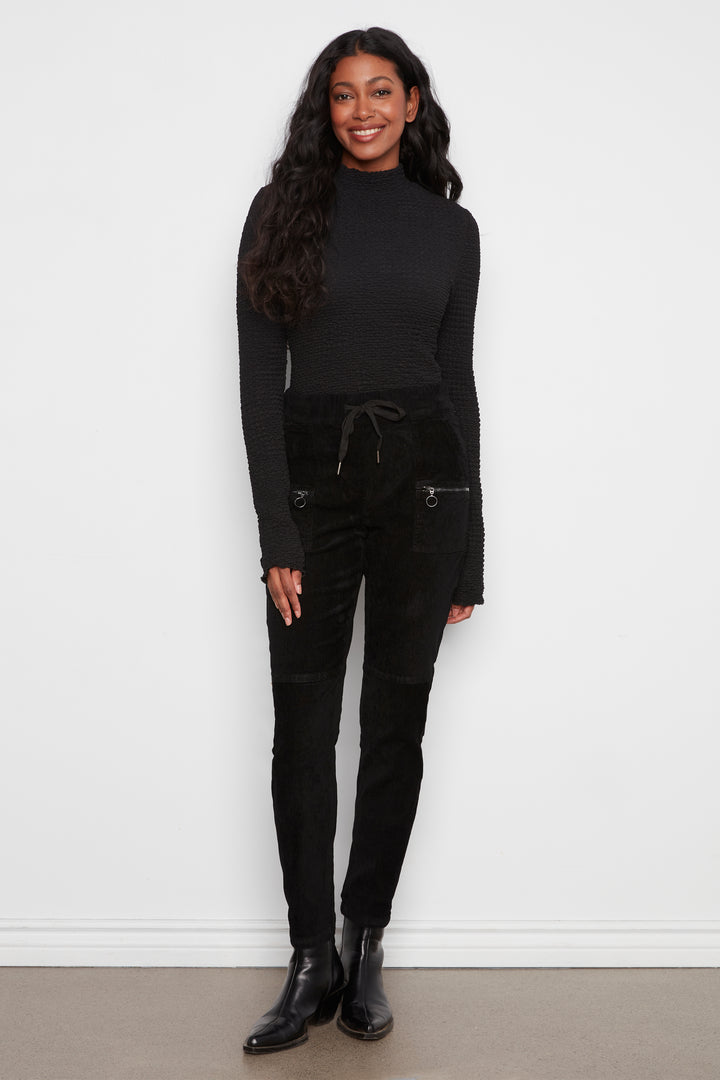 A woman modeling a black ribbed long-sleeve mock neck top and black corduroy pants with zippered pockets, standing against a plain white wall.