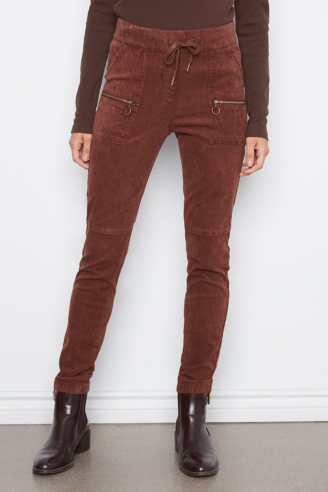 A person wearing brown corduroy jogger-style pants with a drawstring waist and zippered side pockets, paired with a brown long-sleeve top and dark brown ankle boots, standing against a white wall.