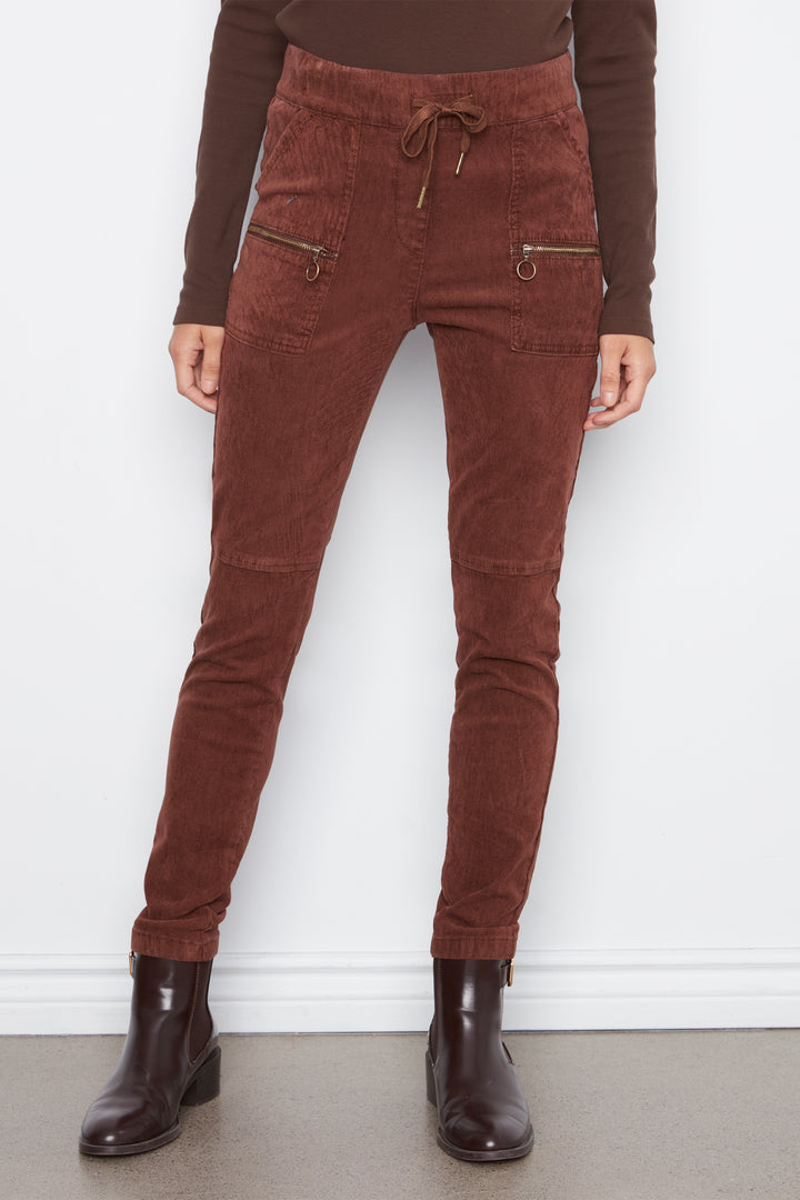 A person wearing brown corduroy jogger-style pants with a drawstring waist and zippered side pockets, paired with a brown long-sleeve top and dark brown ankle boots, standing against a white wall.