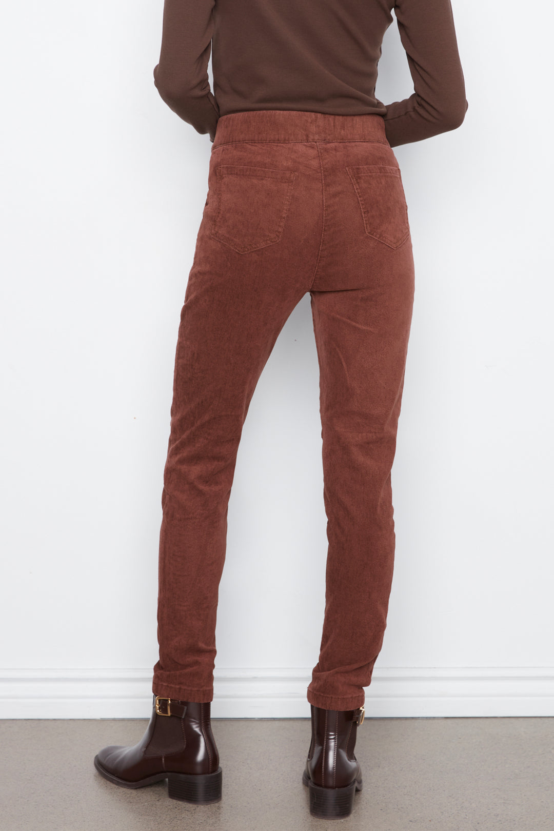 Rear view of a person wearing rust-brown corduroy pants, a dark brown long-sleeve top, and dark brown ankle boots against a white wall.