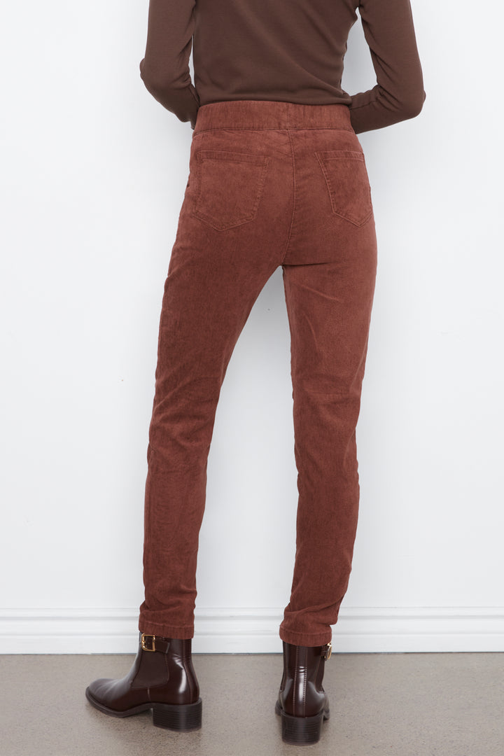 Rear view of a person wearing rust-brown corduroy pants, a dark brown long-sleeve top, and dark brown ankle boots against a white wall.