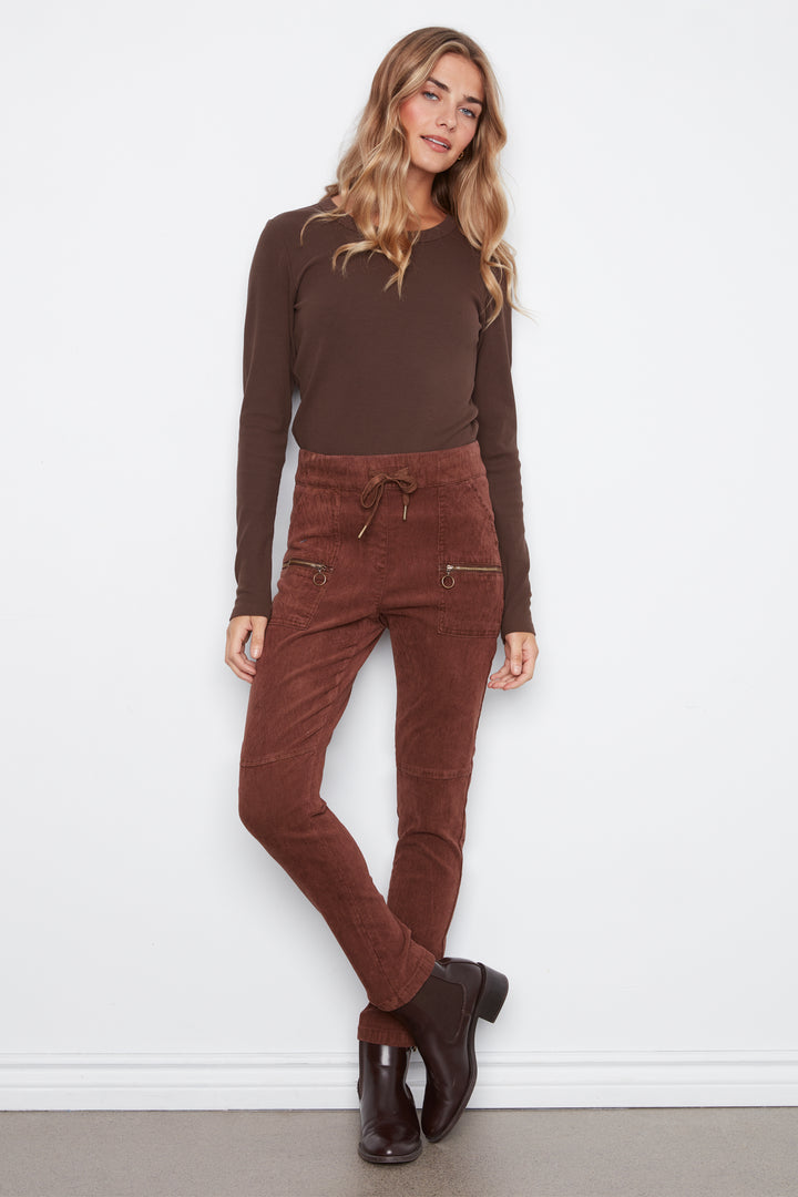 A woman modeling a dark brown long-sleeve knit top and rust-brown corduroy pants with zippered pockets, paired with dark brown leather boots, against a white wall.