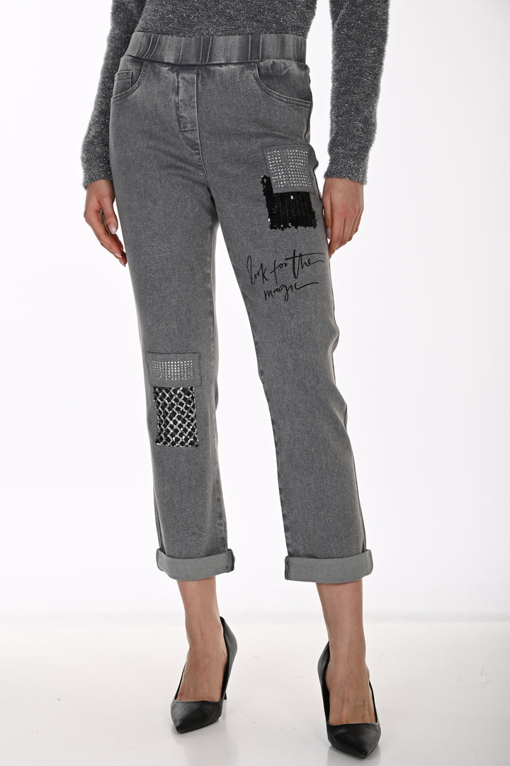 A model wearing gray patchwork cropped jeans with black and white patterned pockets, a black graphic patch, and script embroidery on the thigh, paired with a gray long-sleeve top and black heels.