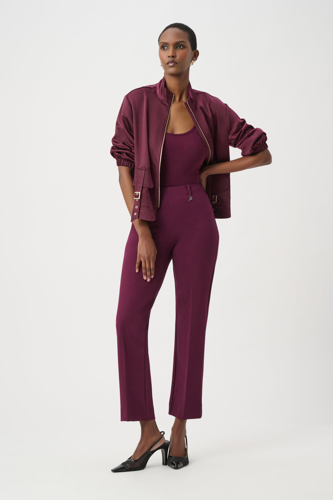 A model wearing a matching burgundy three-piece set consisting of a bomber jacket, a scoop-neck top, and cropped trousers, paired with black pointed-toe heels against a plain white studio background.