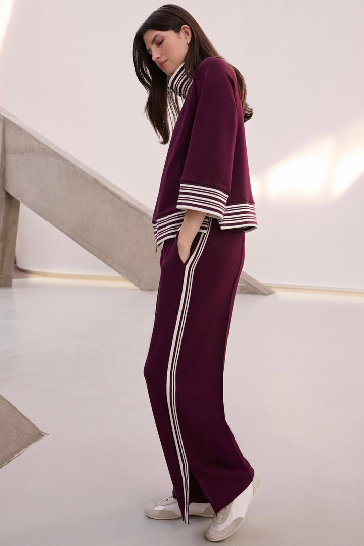 A woman modeling a matching deep burgundy knit lounge set with white and navy striped accents on the sleeves and side stripes on the pants, standing in a minimalist modern interior.