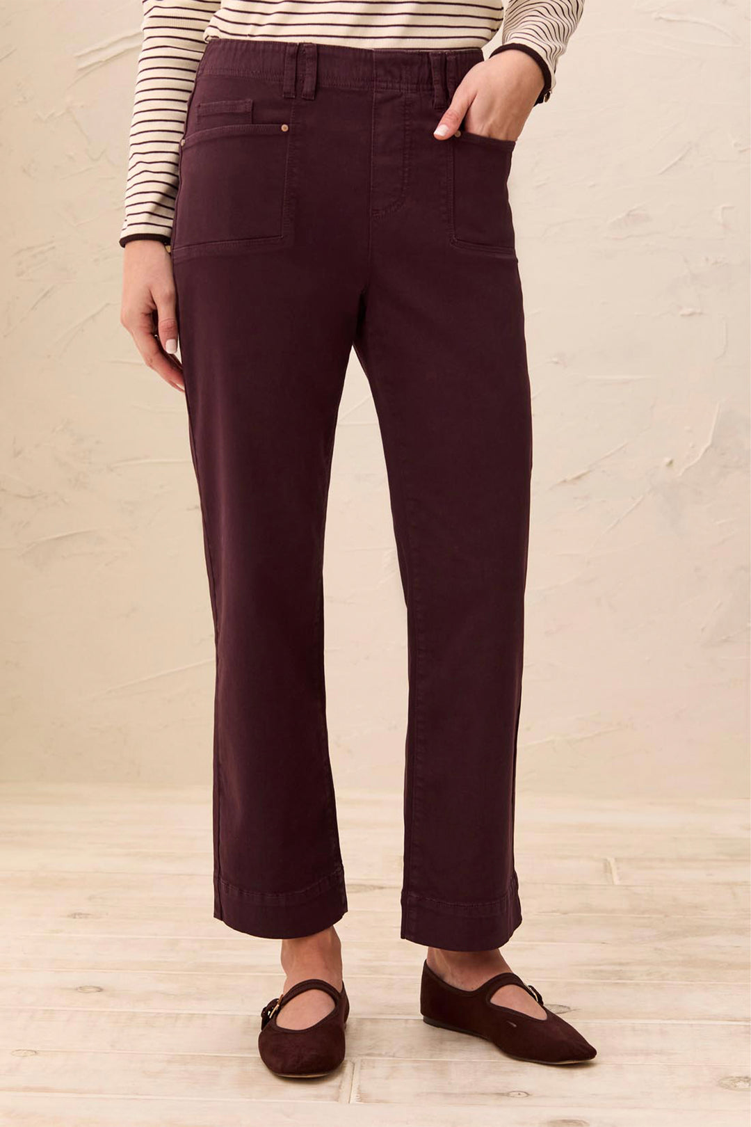 A person wearing dark plum-colored straight-leg pants, a striped long-sleeve top, and dark brown flat shoes, standing against a textured beige wall and light wood floor.