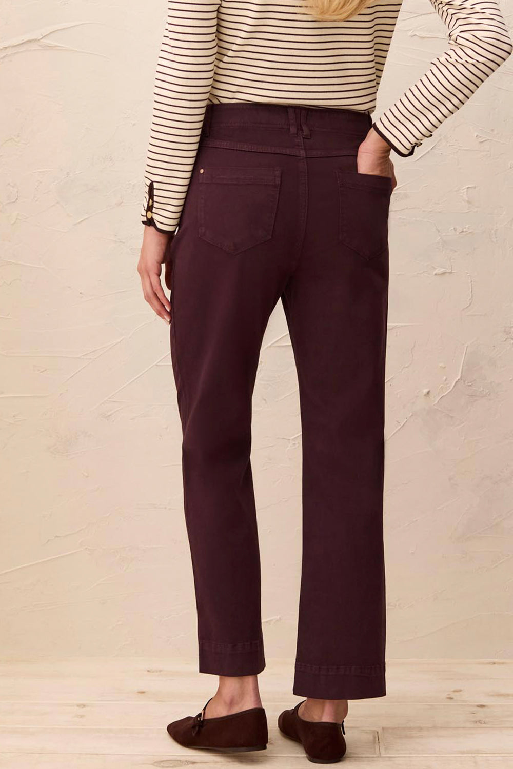 A person shown from the back wearing dark plum-colored straight-leg pants, a cream and brown striped long-sleeve top, and brown loafers, standing against a textured beige wall.