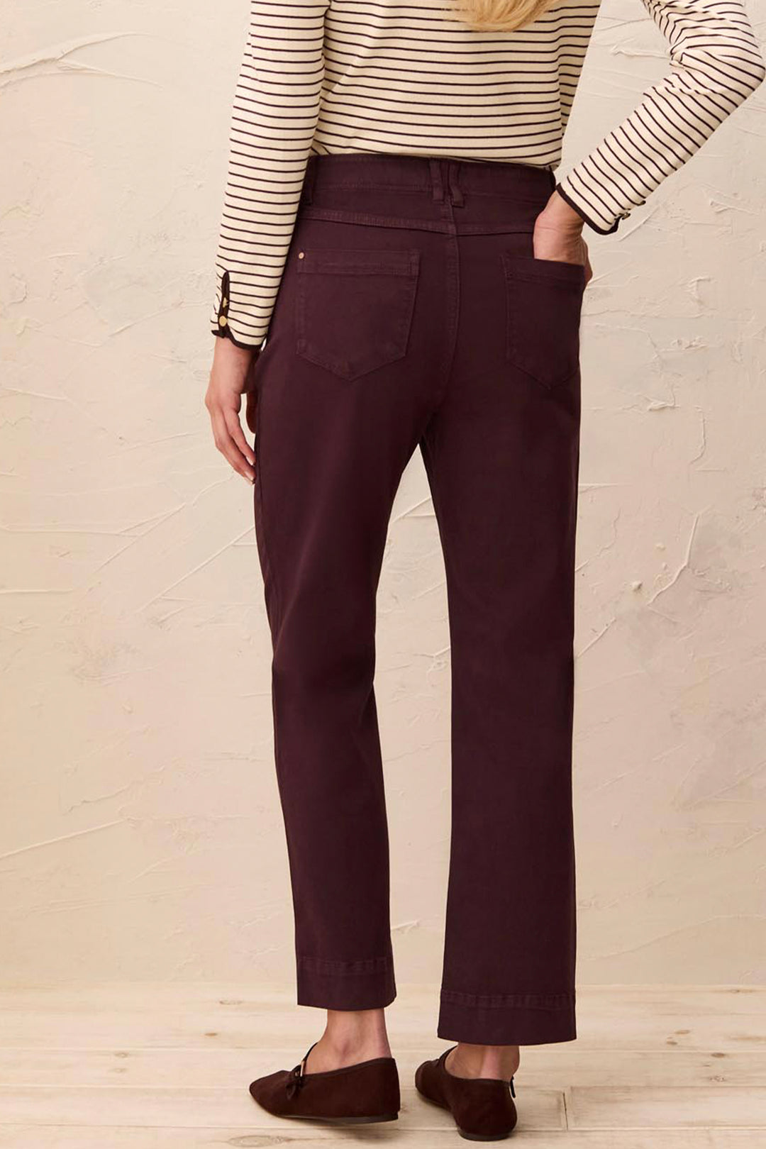 A person shown from the back wearing dark plum-colored straight-leg pants, a cream and brown striped long-sleeve top, and brown loafers, standing against a textured beige wall.