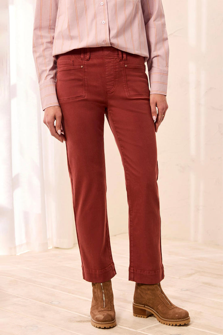 A person wearing rust-red straight-leg pants, a light pink striped button-up shirt, and brown ankle boots, standing in a softly lit indoor setting with light wood flooring and white curtains.