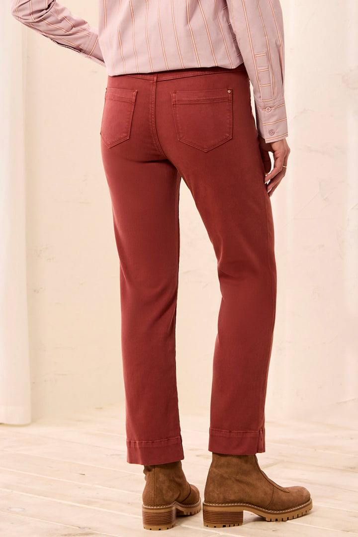 A person shown from the back wearing rust-red straight-leg pants, a light pink striped shirt, and brown suede boots in a bright indoor setting.