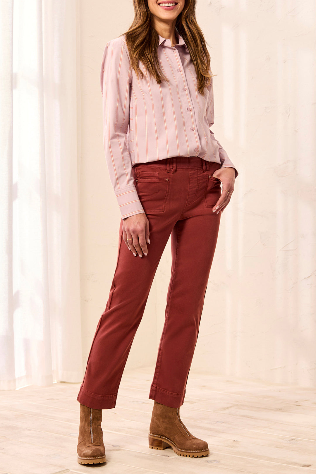A woman modeling a light pink striped button-down shirt and rust-red straight-leg jeans with brown ankle boots, standing in a bright indoor setting with white curtains.