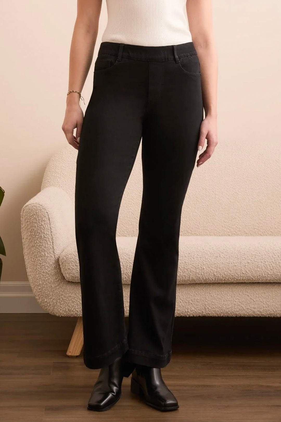 A person wearing black wide-leg cropped pants with a white ribbed top and black ankle boots, standing in front of a cream boucle sofa against a beige wall.