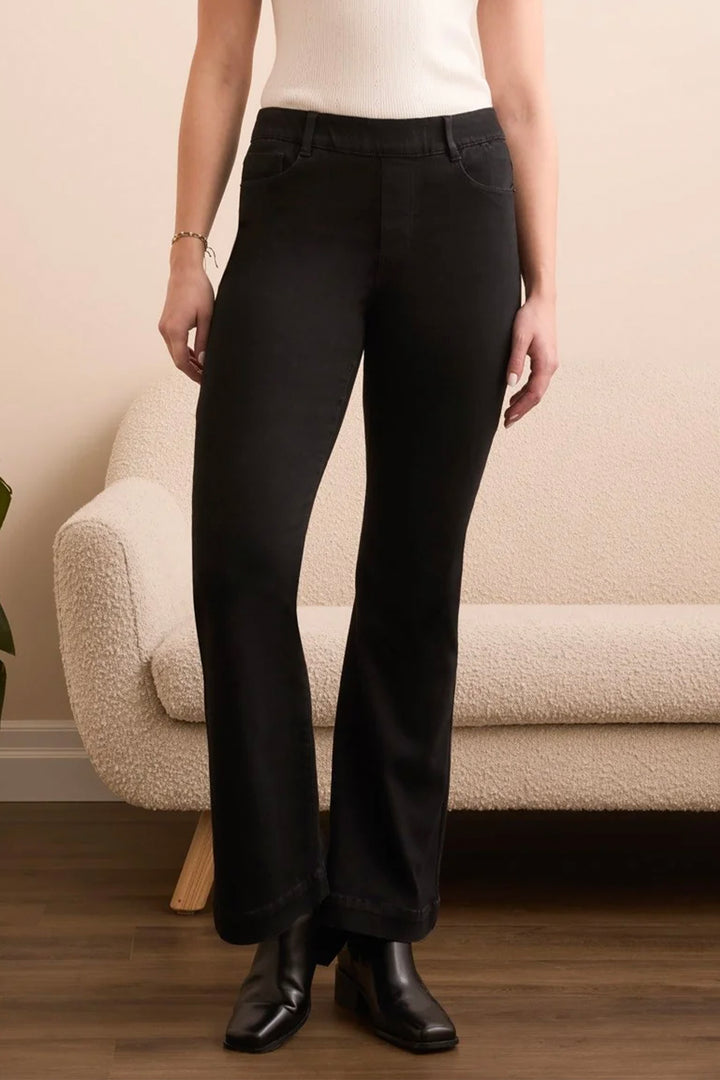 A person wearing black wide-leg cropped pants with a white ribbed top and black ankle boots, standing in front of a cream boucle sofa against a beige wall.