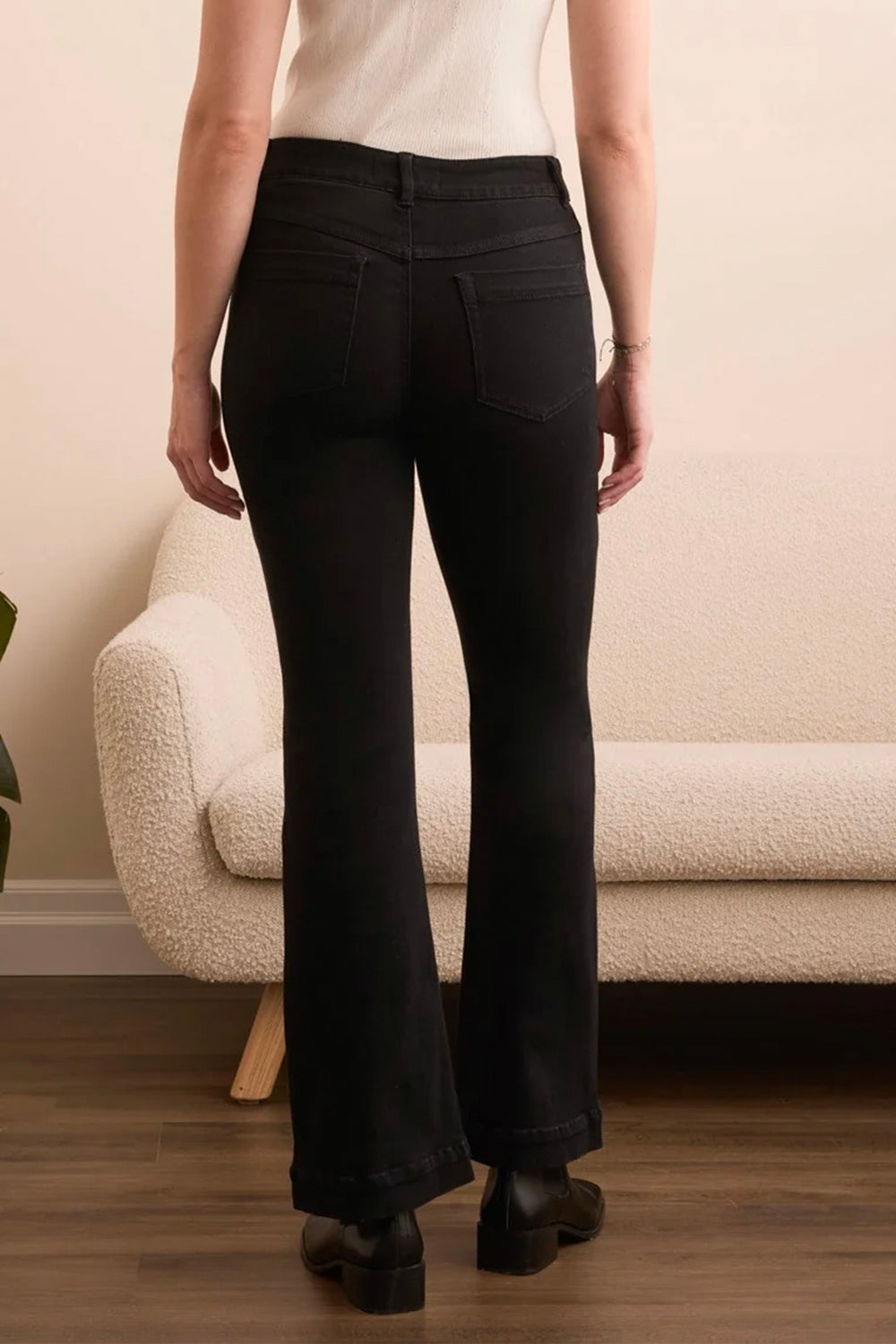 A person shown from the back wearing black flared jeans, standing in a room with a cream boucle sofa and wood floor.