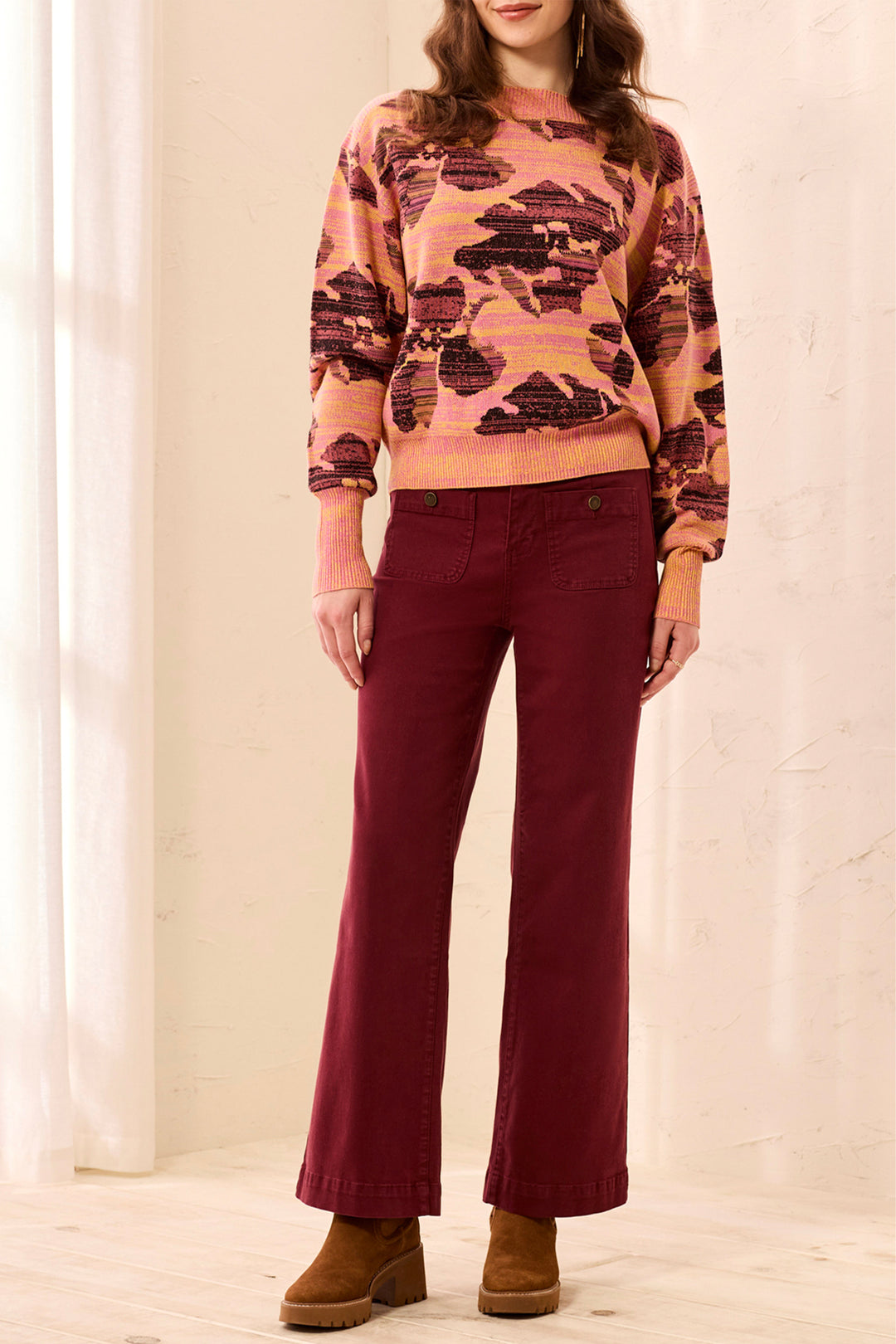 A person wearing burgundy straight-leg pants, a pink and brown patterned sweater, and brown block-heel mules in a bright indoor setting.
