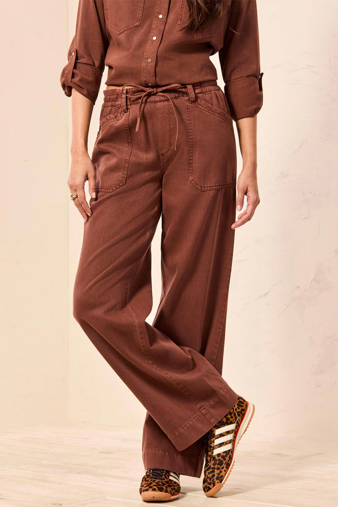 A person wearing rust-brown wide-leg drawstring pants, a matching long-sleeve button-up shirt, and leopard-print sneakers, standing against a light beige wall.
