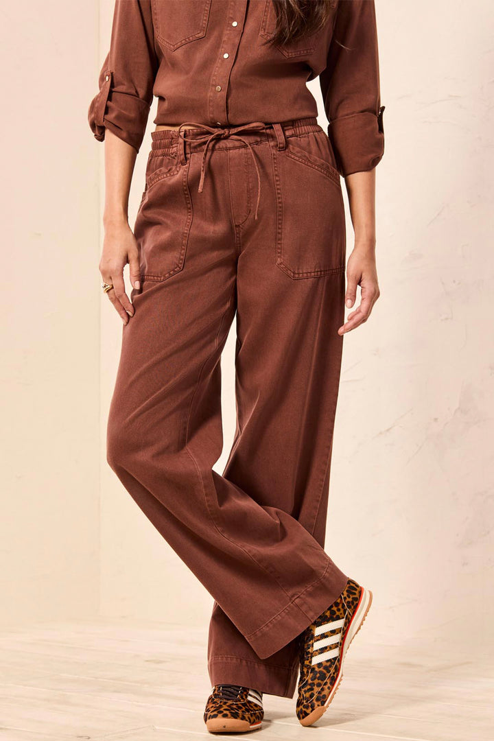 A person wearing rust-brown wide-leg drawstring pants, a matching long-sleeve button-up shirt, and leopard-print sneakers, standing against a light beige wall.