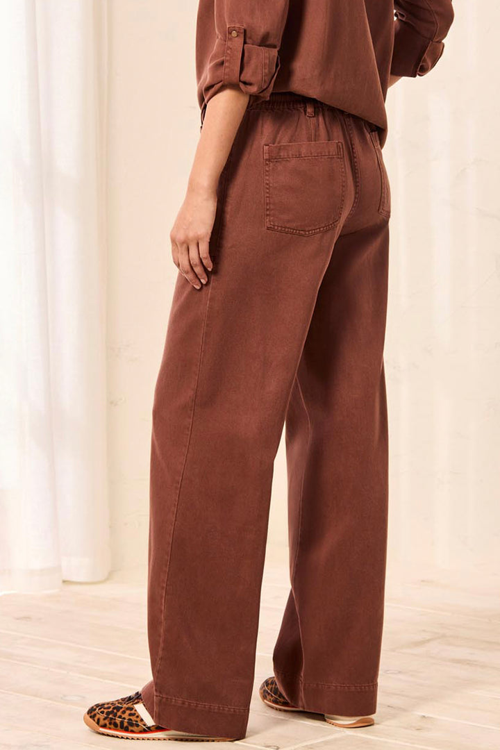 A person shown from the back wearing rust-brown wide-leg pants, a matching rust-brown top, and leopard-print flat shoes in a bright indoor setting with light curtains and a pale wood floor.