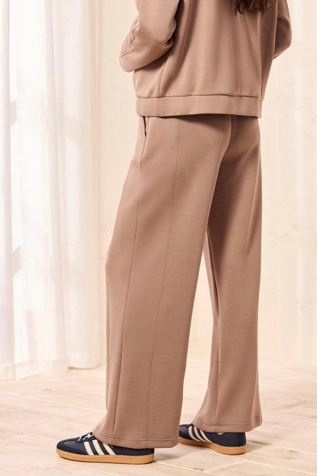 A person wearing a matching taupe-brown lounge set with wide-leg pants and a cropped sweatshirt, paired with navy sneakers, standing in a bright room with sheer curtains.
