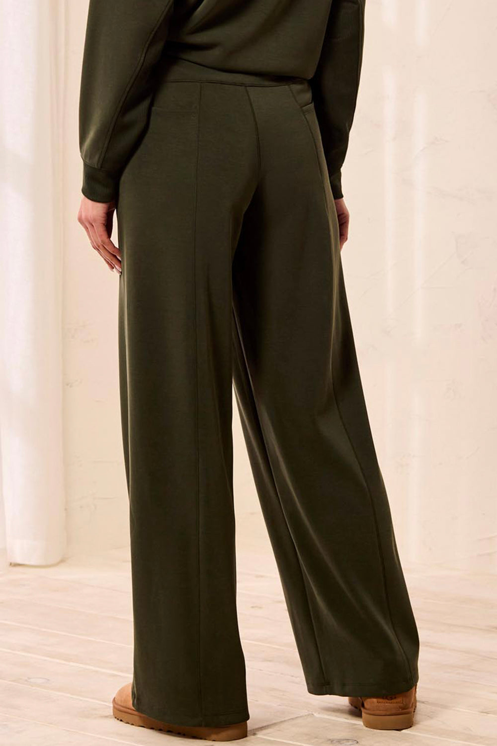 A person shown from the back wearing dark olive green wide-leg trousers and a matching long-sleeve top, standing in a bright indoor setting with light wood flooring.