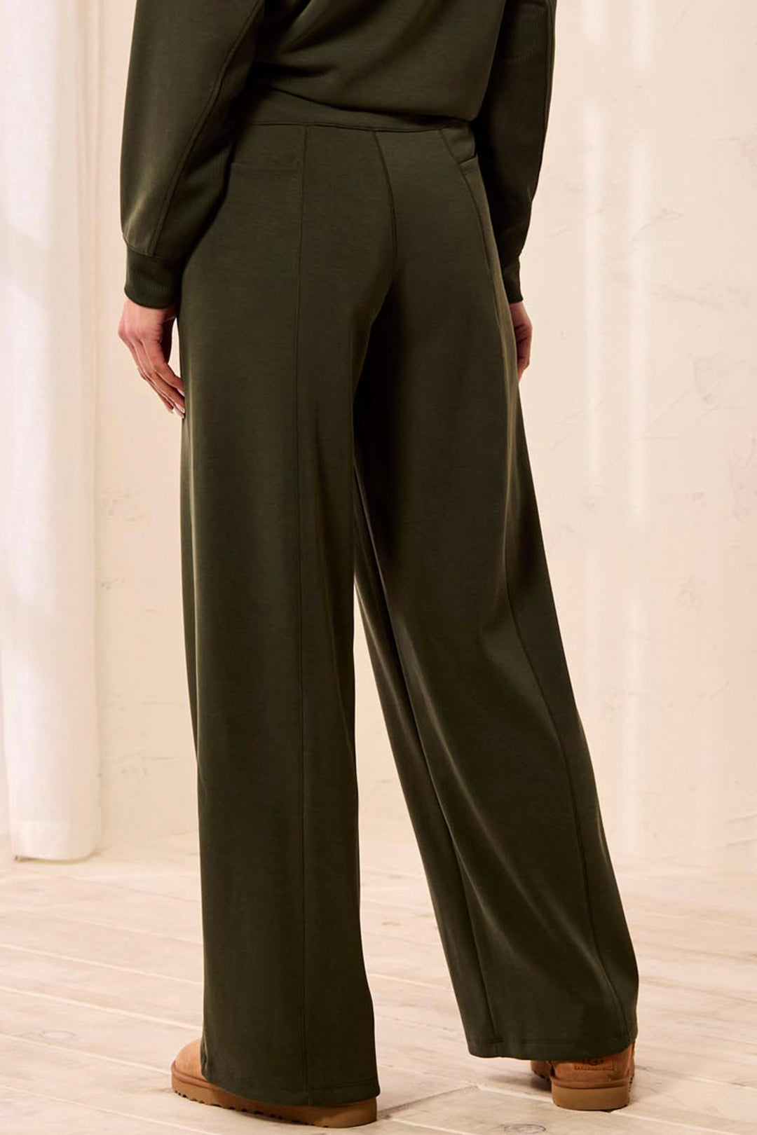 A person shown from the back wearing dark olive green wide-leg trousers and a matching long-sleeve top, standing in a bright indoor setting with light wood flooring.