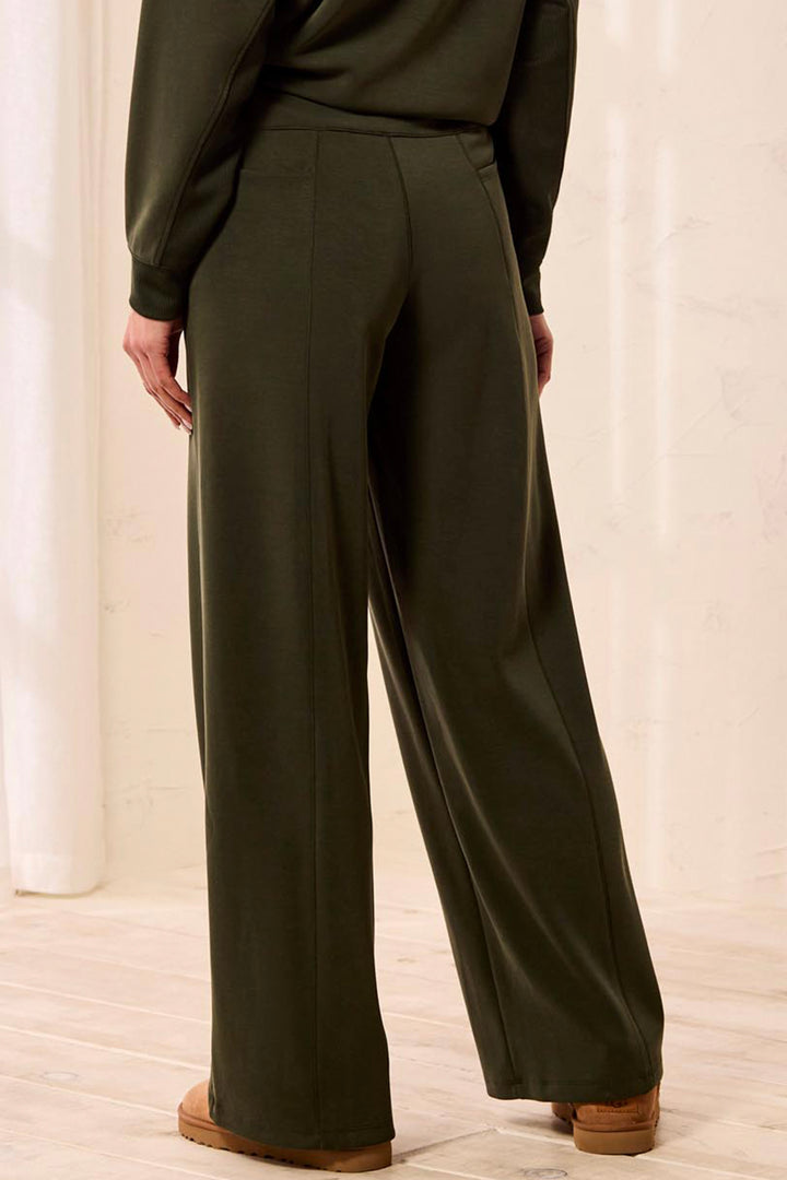 A person shown from the back wearing dark olive green wide-leg trousers and a matching long-sleeve top, standing in a bright indoor setting with light wood flooring.