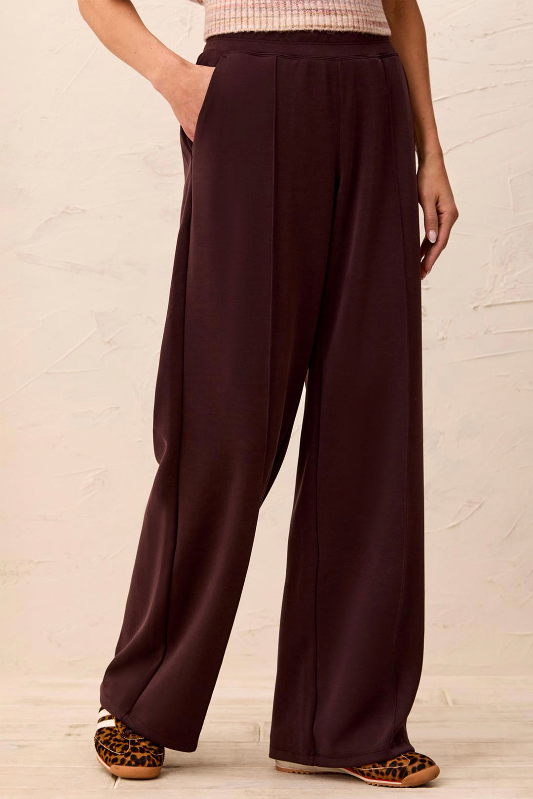 A person wearing dark plum wide-leg pants with a textured cream top and leopard-print shoes, standing against a light textured wall.