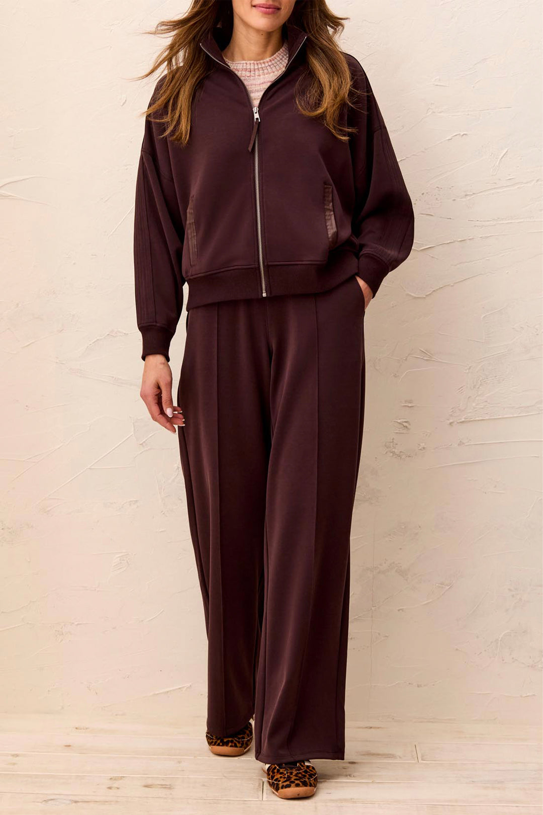 A woman modeling a matching two-piece lounge set in deep plum purple, featuring a zip-up jacket and wide-leg pants, styled with a pink knit top and leopard-print slippers against a textured beige wall.