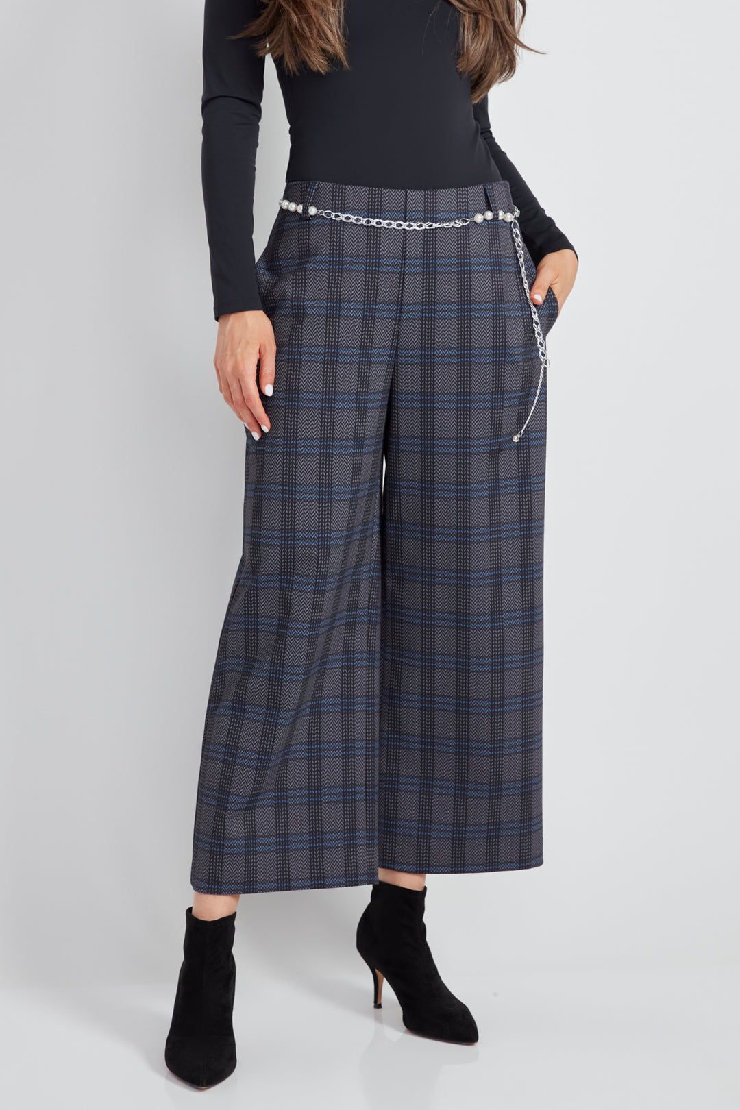 A woman standing in front of a light grey background wearing a long sleeve top with plaid wide leg crop pants.  