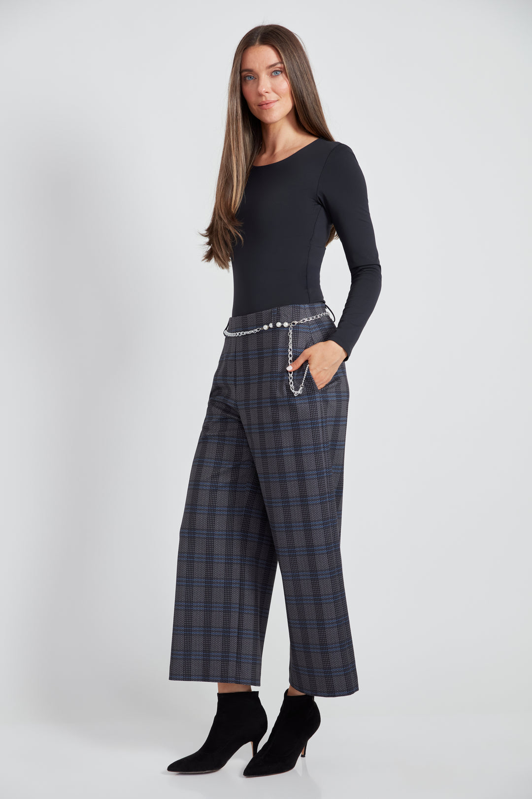 A woman standing in front of a light grey background wearing a long sleeve top with plaid wide leg crop pants.  