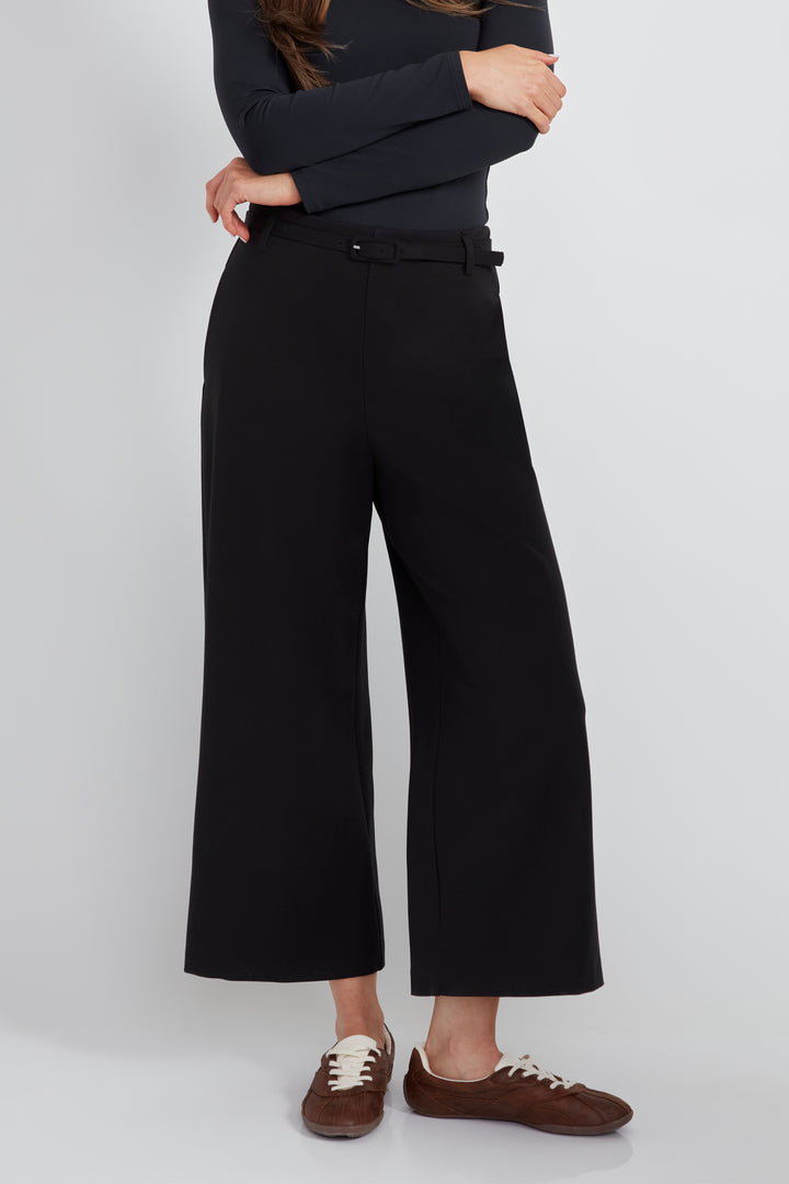 A woman standing in front of a light grey background wearing a long sleeve top and black wide leg pants with matching black belt. 