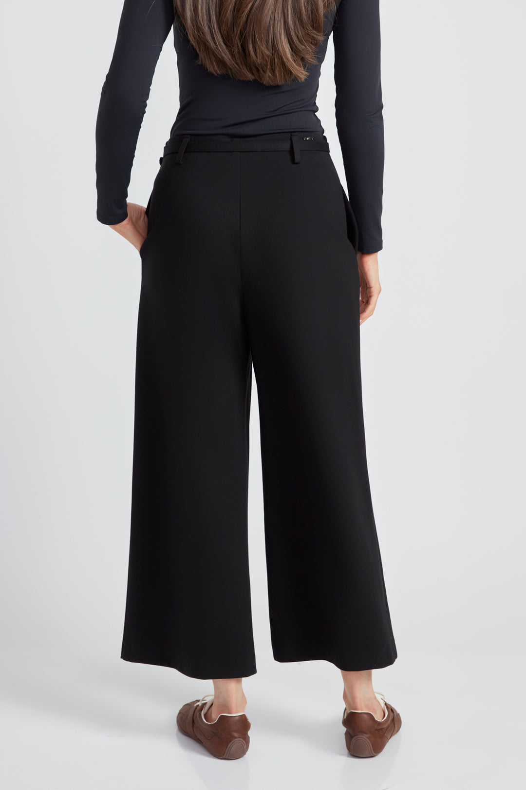 A woman standing in front of a light grey background wearing a long sleeve top and black wide leg pants with matching black belt. 