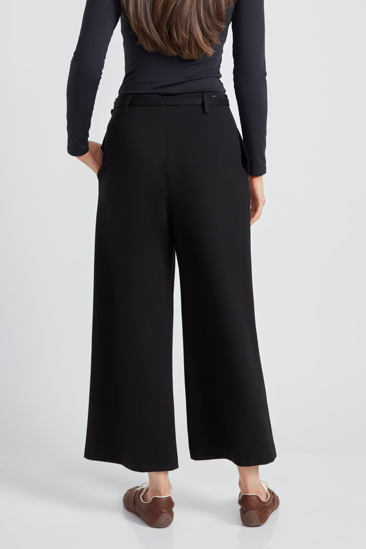 A woman standing in front of a light grey background wearing a long sleeve top and black wide leg pants with matching black belt. 