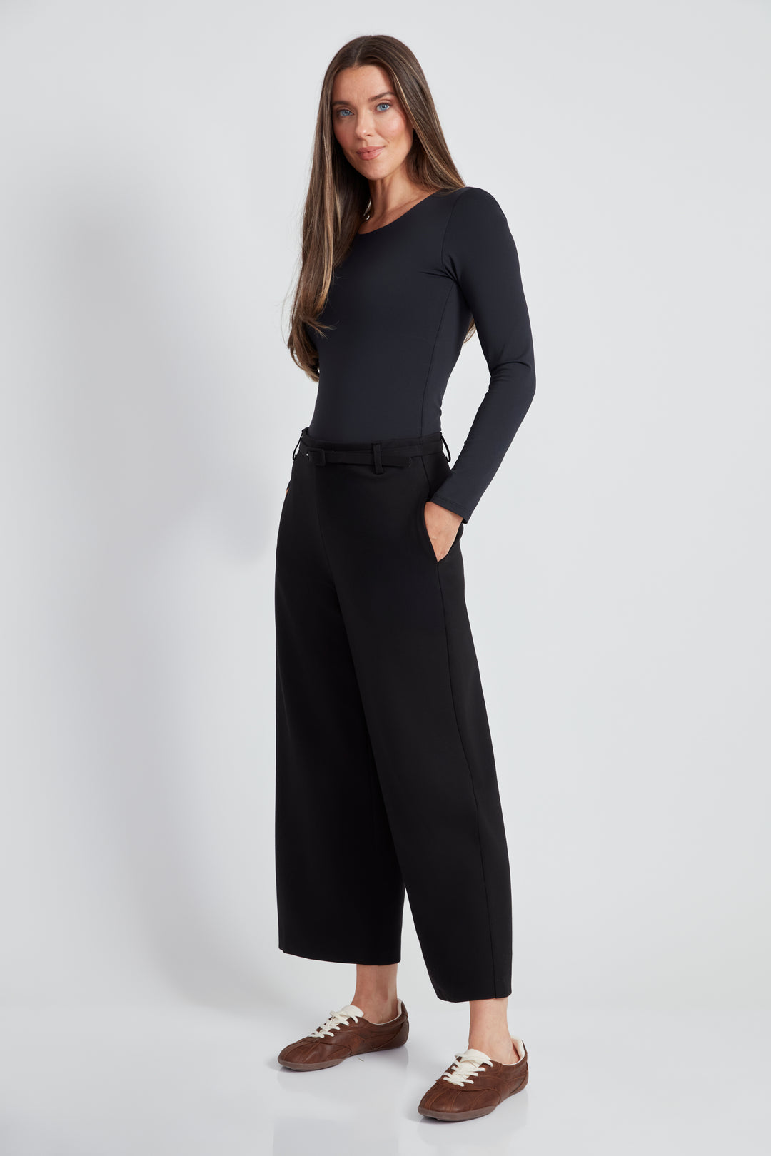 A woman standing in front of a light grey background wearing a long sleeve top and black wide leg pants with matching black belt. 