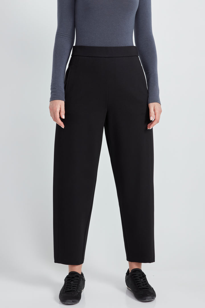 A woman wearing a long sleeve top and black  barrel leg pants standing in front of a light grey background. 