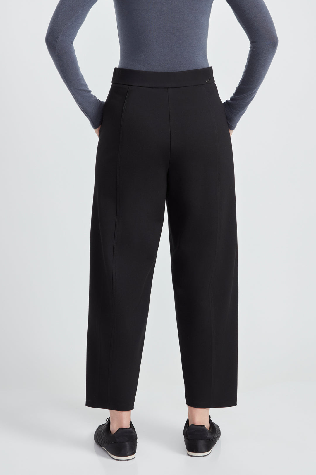 A woman wearing a long sleeve top and black  barrel leg pants standing in front of a light grey background. 