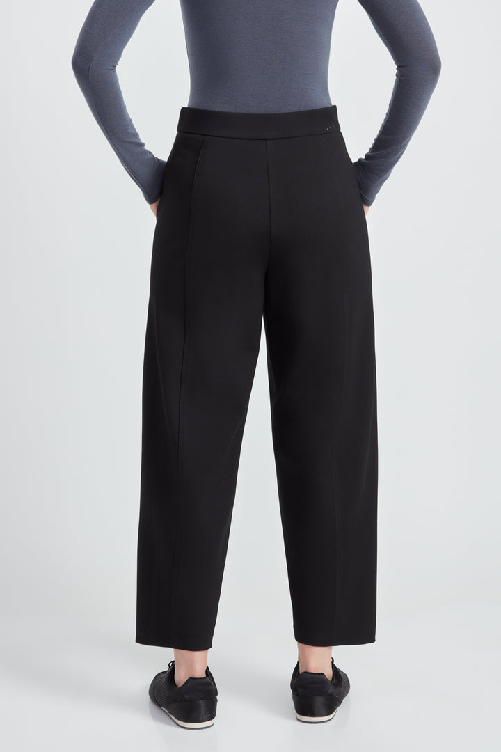 A woman wearing a long sleeve top and black  barrel leg pants standing in front of a light grey background. 