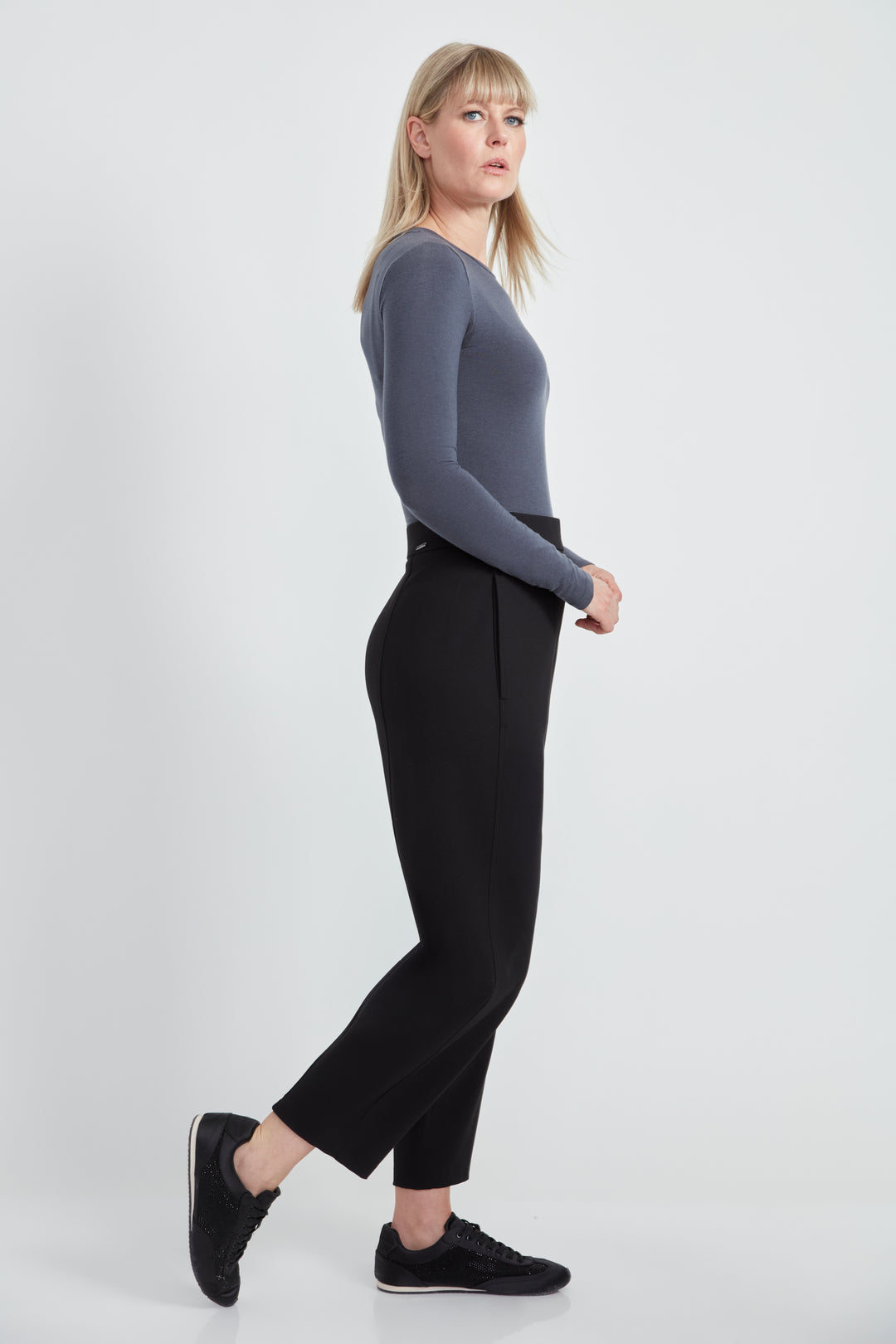 A woman wearing a long sleeve top and black  barrel leg pants standing in front of a light grey background. 