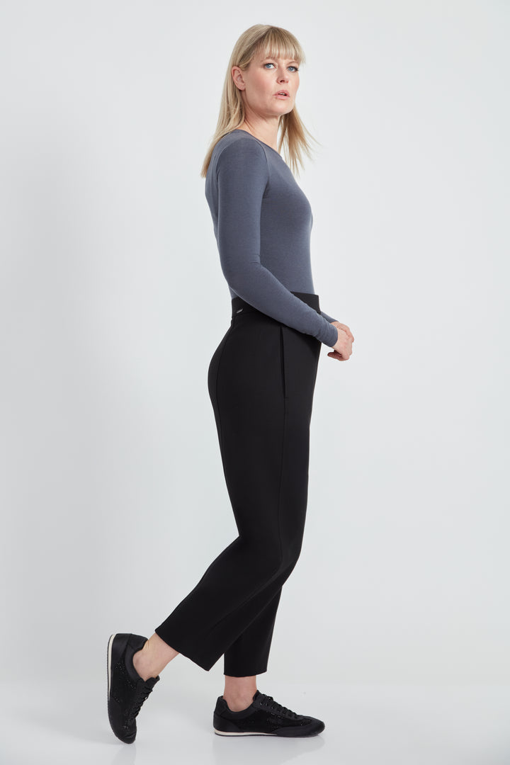 A woman wearing a long sleeve top and black  barrel leg pants standing in front of a light grey background. 