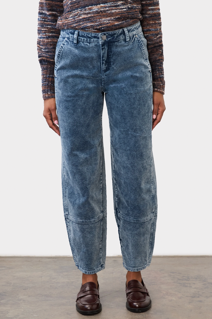 A woman wearing a bulky sweater and light wash corduroy barrel jeans standing in front of a grey studio background. 