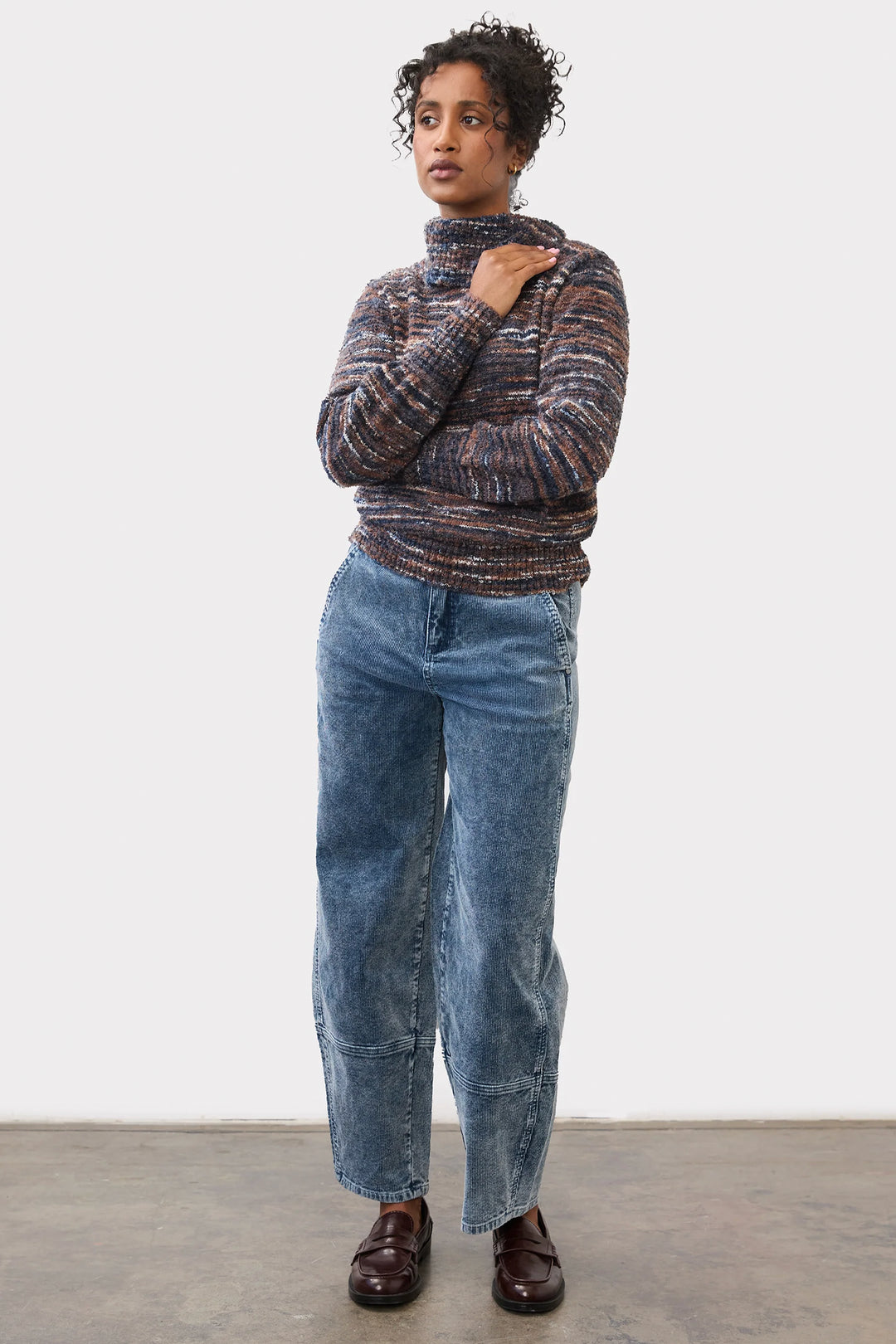 A woman wearing a bulky sweater and light wash corduroy barrel jeans standing in front of a grey studio background. 