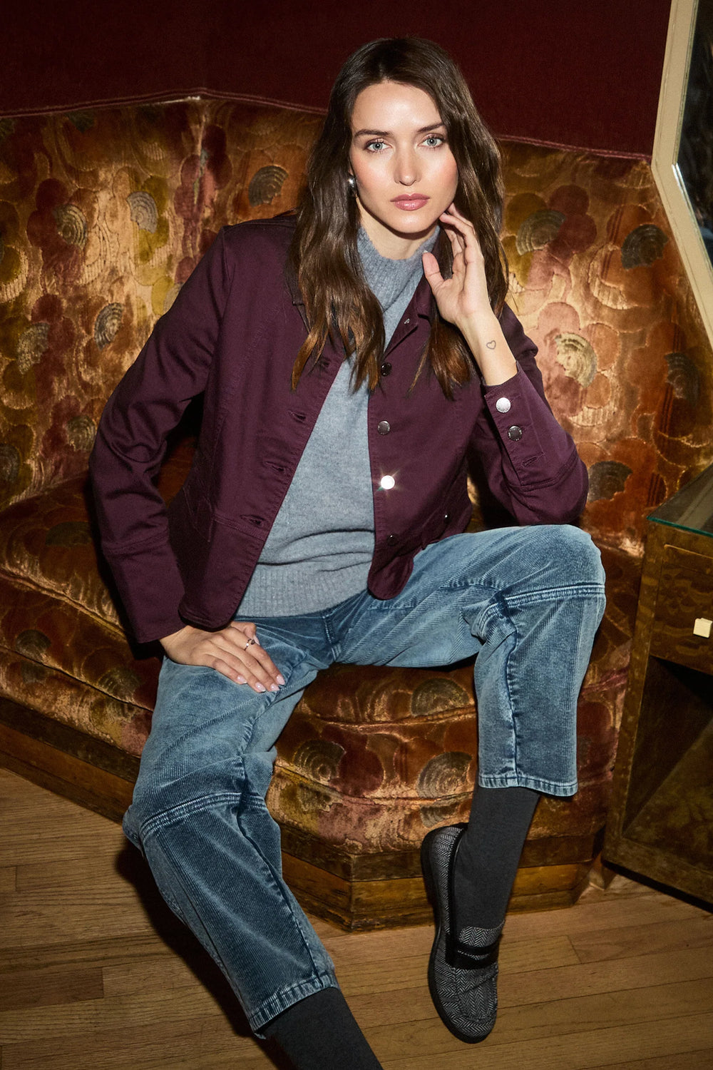 A woman wearing a burgundy jacket=y, grey sweater and light wash corduroy barrel jeans sitting on a printed couch. 