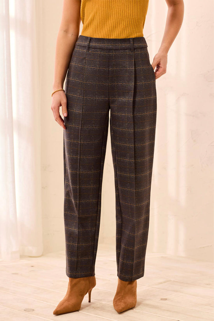 A woman wearing brown plaid trousers standing in front of a beige background. 