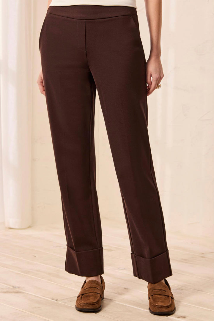 A woman wearing deep brown trousers with a  large cuff standing in front of a beige background. 