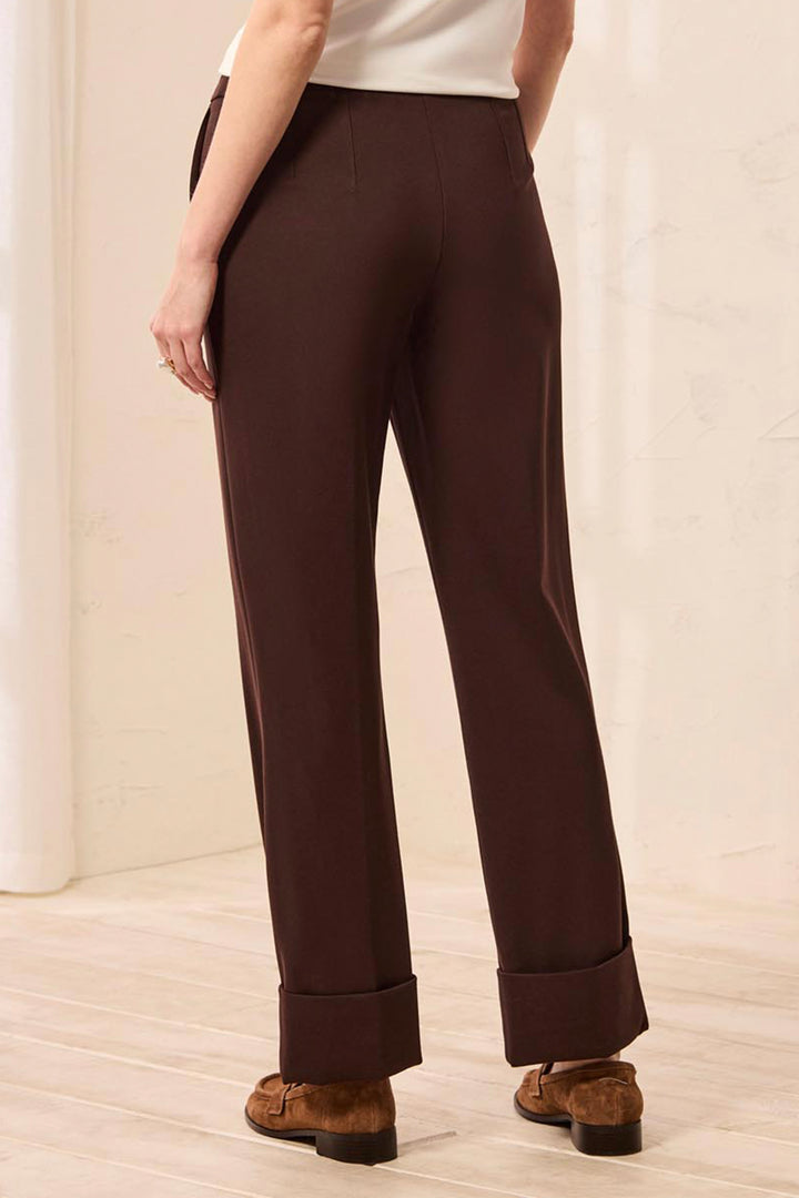 A woman shown from the back wearing deep brown trousers with a  large cuff standing in front of a beige background. 