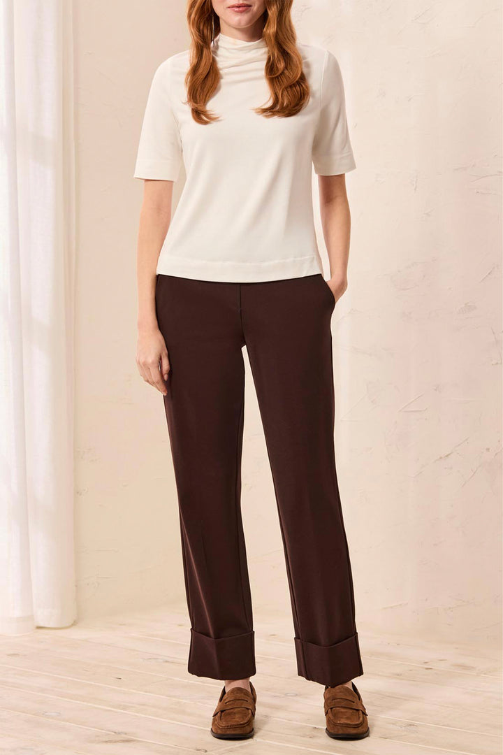 A woman wearing deep brown trousers with a  large cuff standing in front of a beige background. 