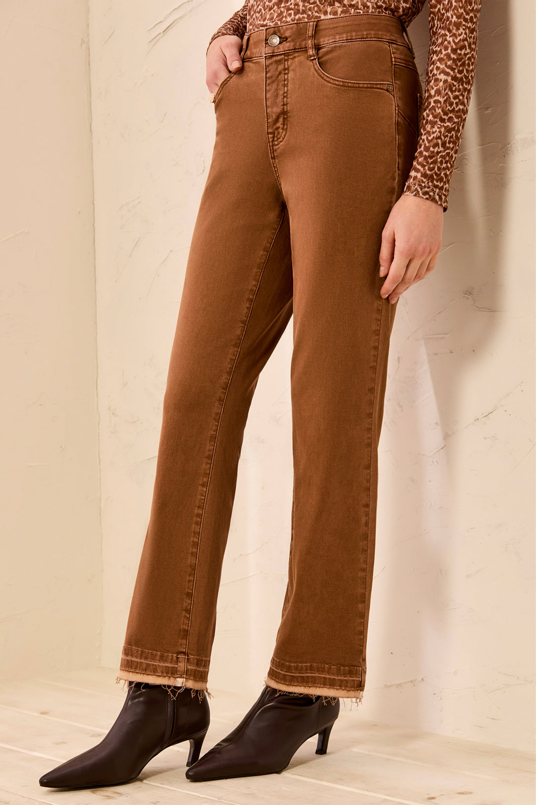 A woman wearing tan pants and brown boots standing in front of a beige background. 