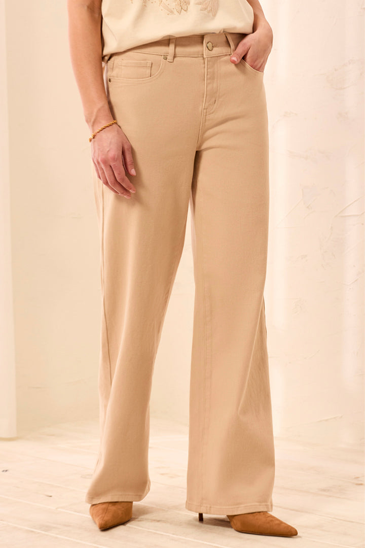 A woman wearing tan wide leg shimmery pants and cream t-shirt standing in front of a beige background. 