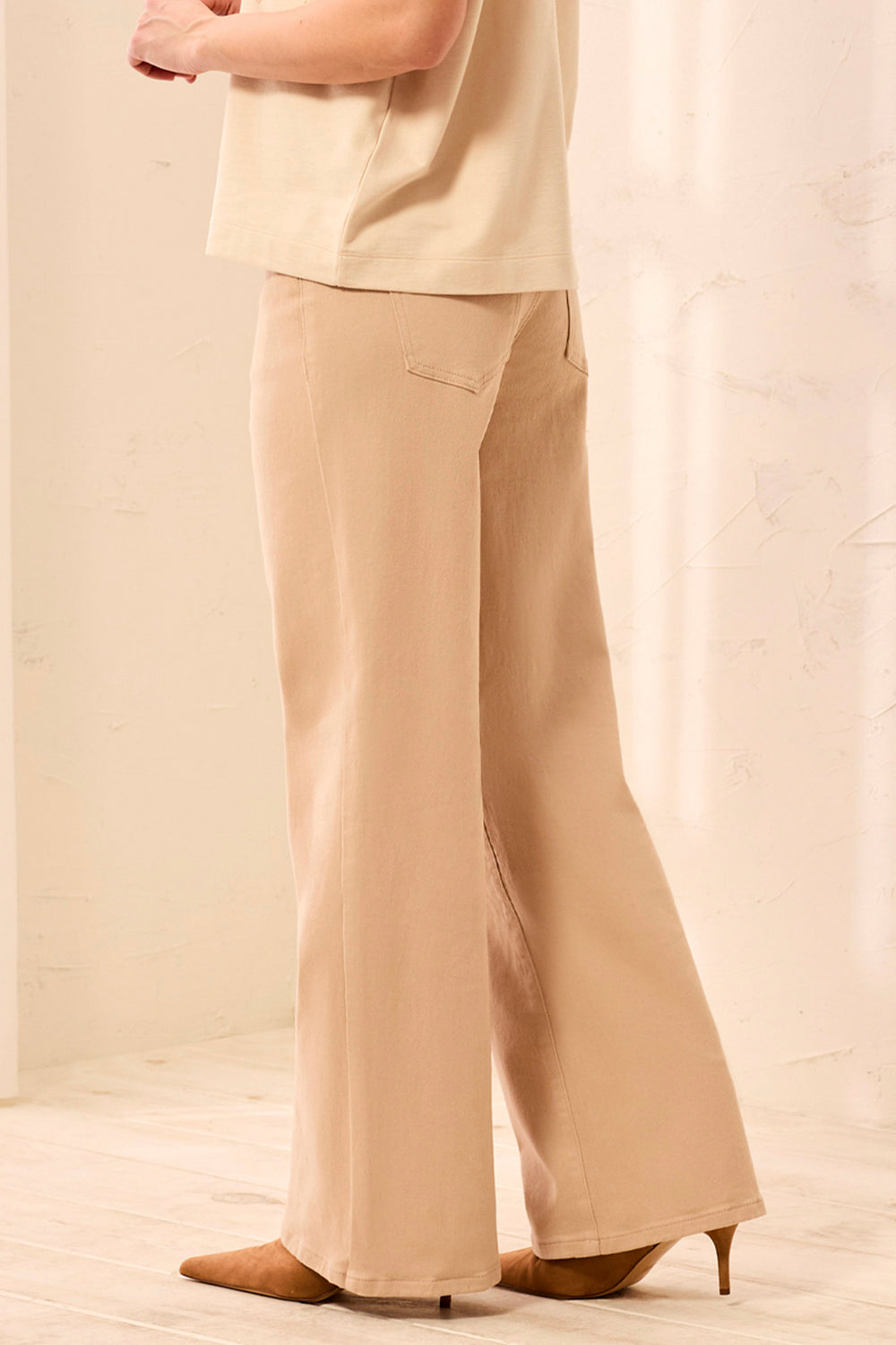 A woman shown from the back wearing tan wide leg shimmery pants and cream t-shirt standing in front of a beige background. 